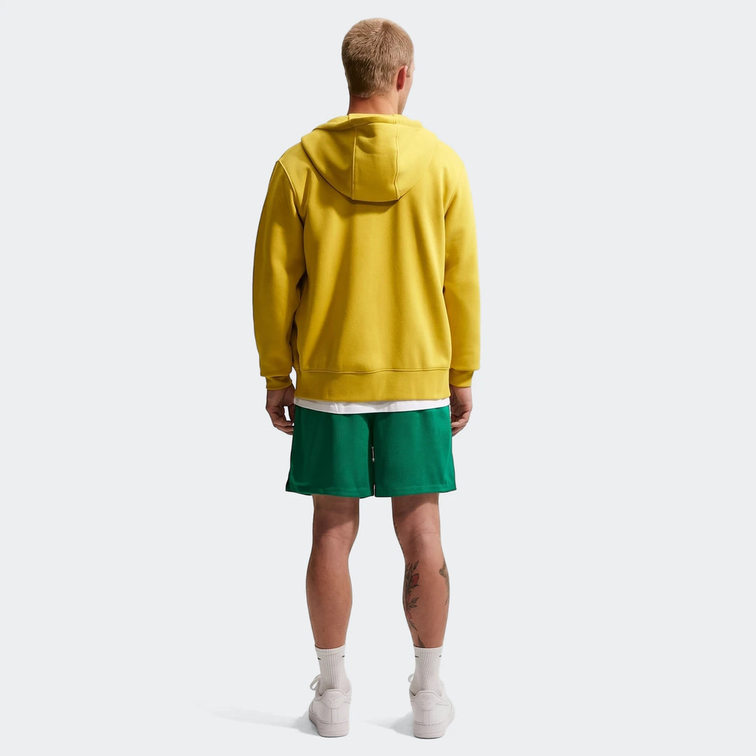Man in Nike Sportswear Club Green Flow Shorts, yellow hoodie, and white sneakers, standing with his back to the camera.