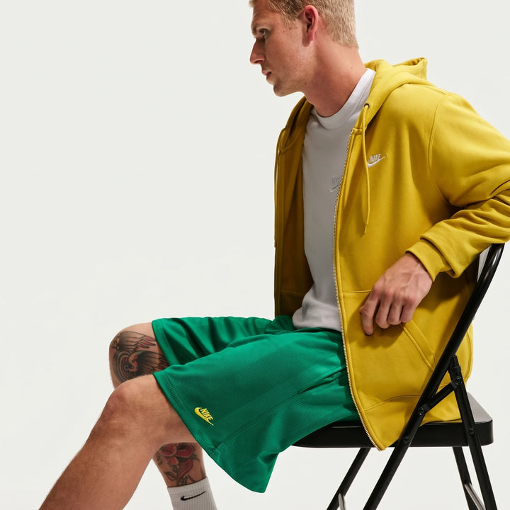Man wearing Nike Sportswear Club green flow shorts and a yellow full-zip hoodie, seated on a black folding chair.
