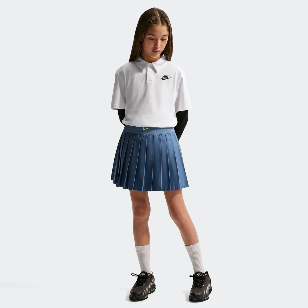 A young girl wears a white Nike Sportswear Club Knit Short-Sleeve Polo, a blue pleated skirt, and black sneakers, captured