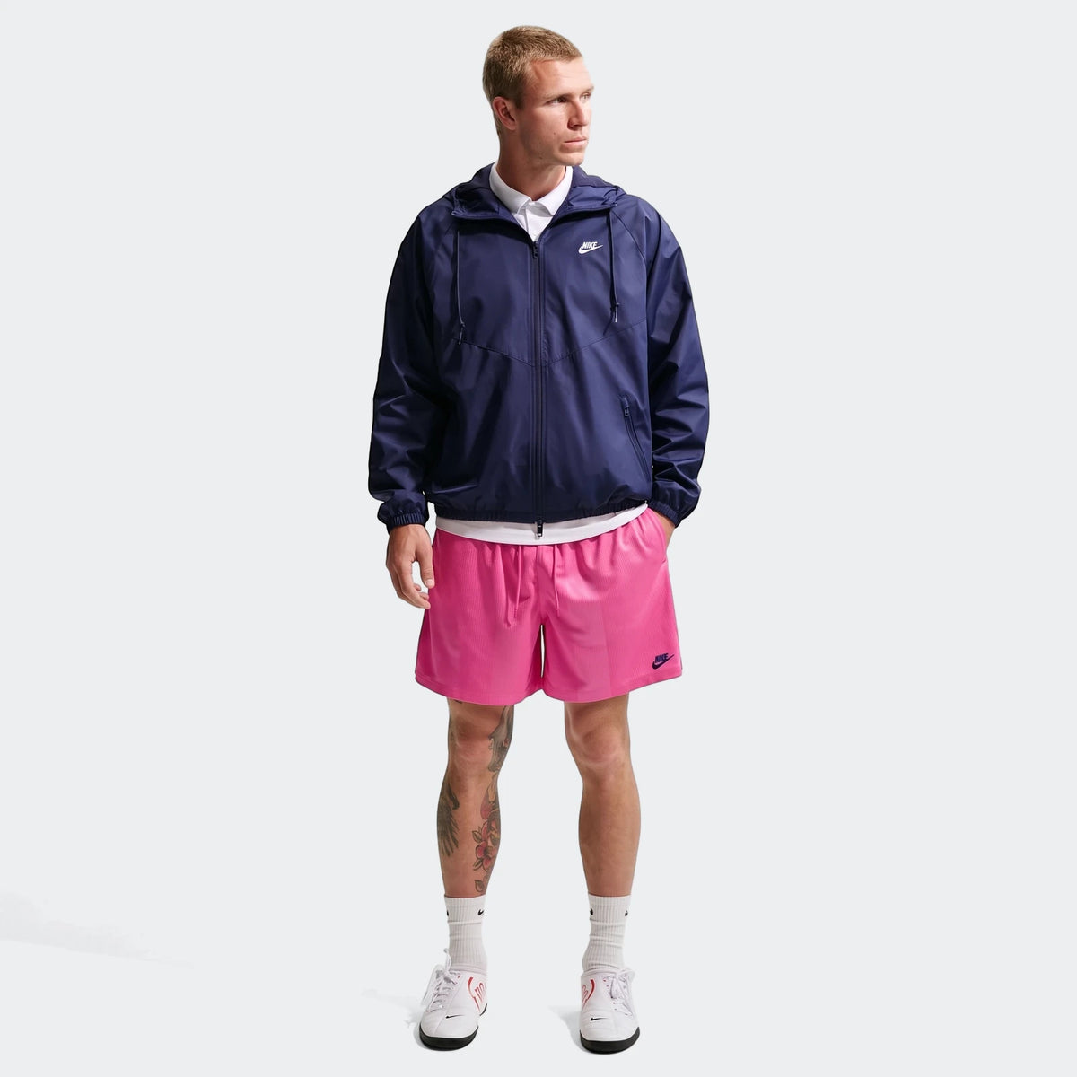 Man standing in full body shot, wearing pink Nike Sportswear Club Flow shorts, a navy Nike windbreaker, and white sneakers.