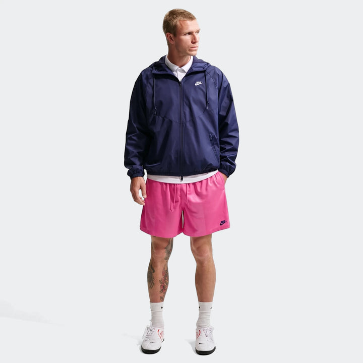 Man standing in full body shot, wearing pink Nike Sportswear Club Flow shorts, a navy Nike windbreaker, and white sneakers.