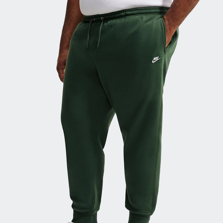 Dark green Nike Sportswear Club men's joggers, featuring a white swoosh logo, shown worn by a person with a hand in the