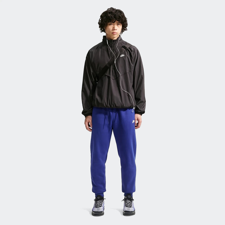 Full body shot of a man wearing blue Nike Sportswear Club Men's Joggers, a black windbreaker, and sneakers, standing against