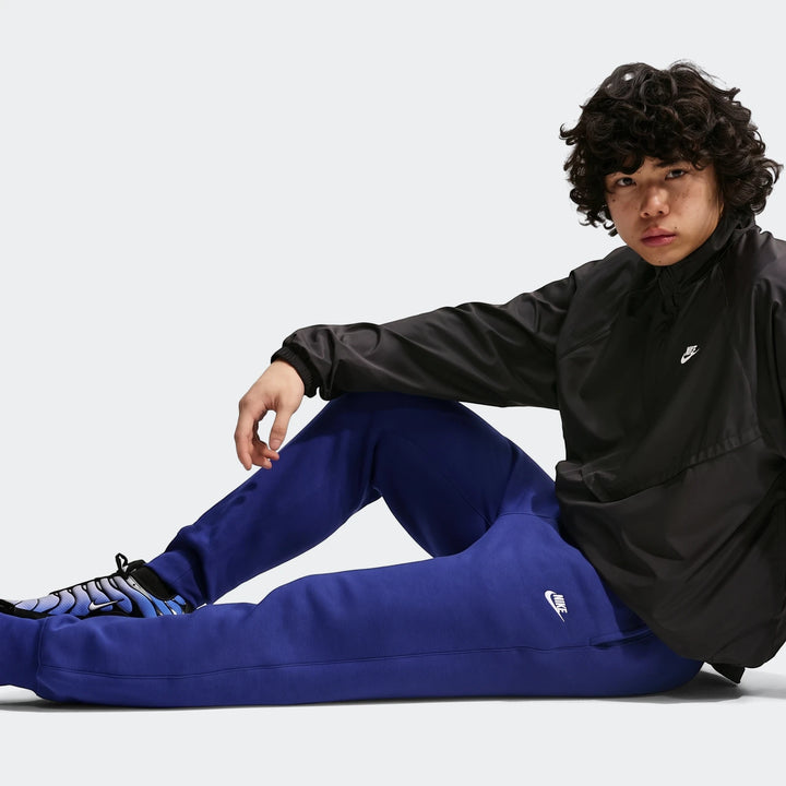 Young man wearing blue Nike Sportswear Club Men's Joggers, paired with a black Nike jacket and blue sneakers, sitting in a