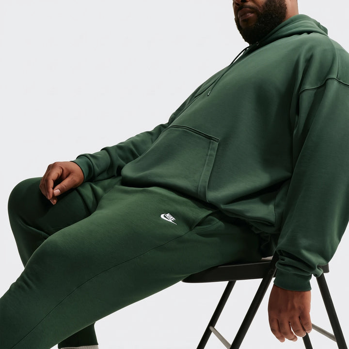 A man wearing dark green Nike Sportswear Club joggers and a matching hoodie, sitting casually on a black stool in a studio