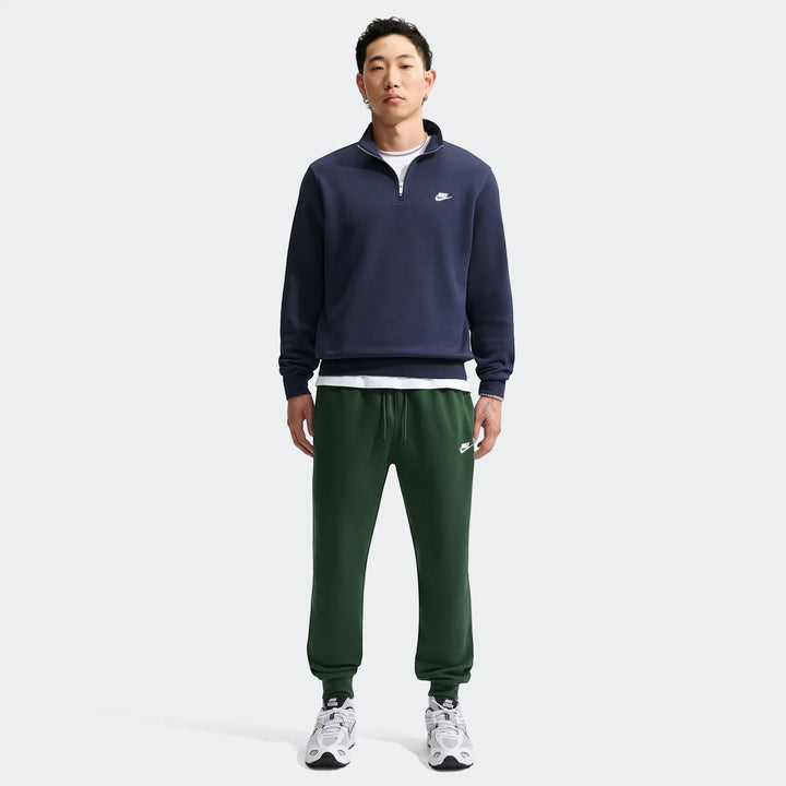 Asian male model wearing dark green Nike Sportswear Club Men's Joggers, a navy half-zip sweatshirt, and white sneakers.