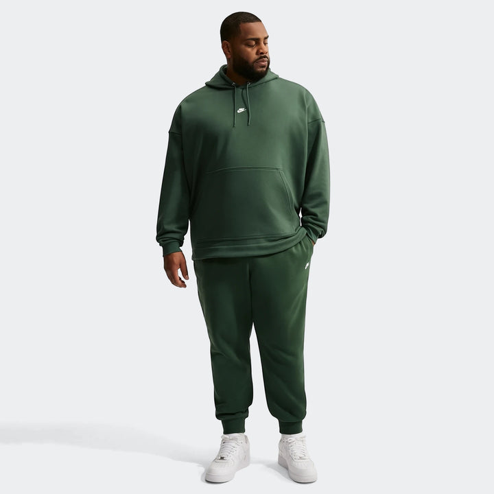 Plus size man wearing a dark green Nike Sportswear Club hoodie and matching joggers with white sneakers.