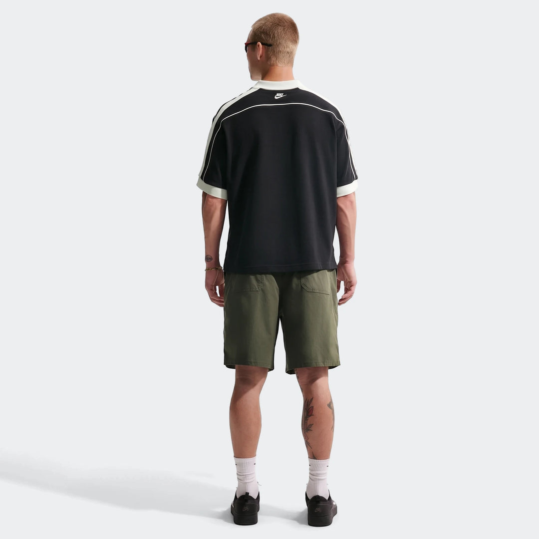 Rear view of a man wearing a Nike Sportswear Club black oversized short-sleeve polo, olive shorts, and black sneakers.