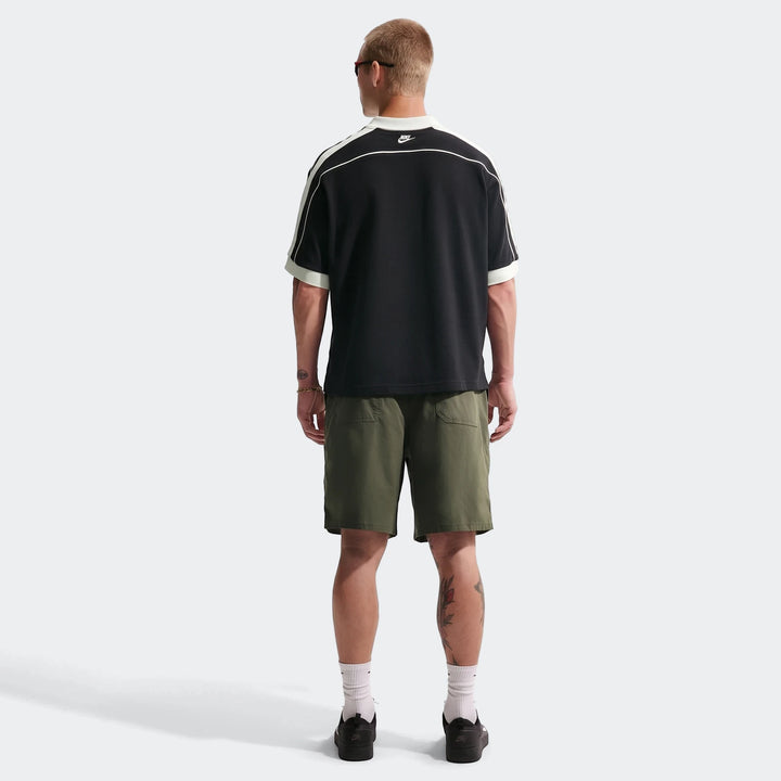 Rear view of a man wearing a Nike Sportswear Club black oversized short-sleeve polo, olive shorts, and black sneakers.