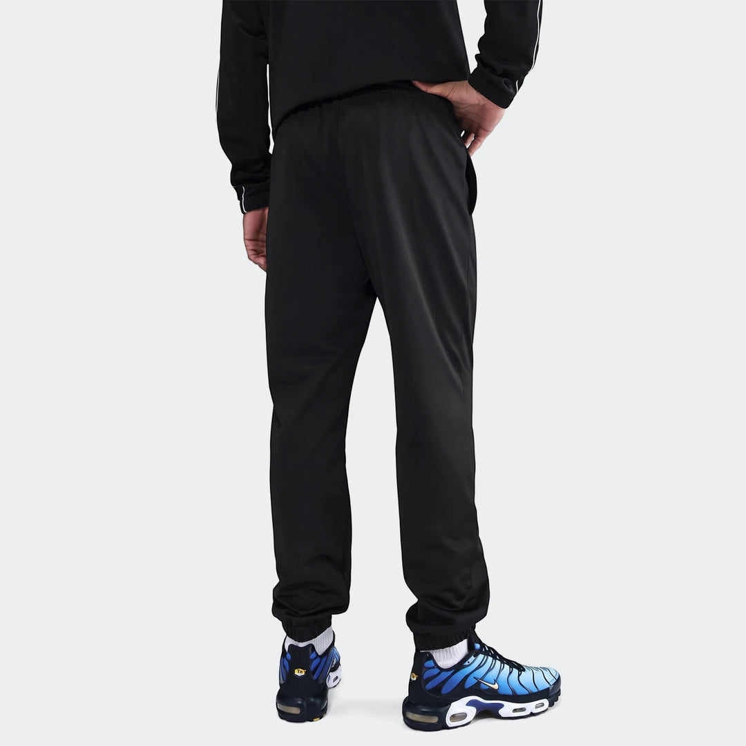 Rear view of a man wearing a black Nike Sportswear Club poly-knit tracksuit and blue Nike sneakers on a white background.