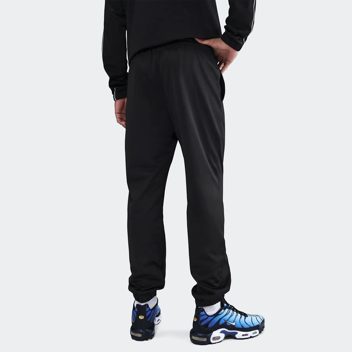 Rear view of a man wearing a black Nike Sportswear Club poly-knit tracksuit and blue Nike sneakers on a white background.