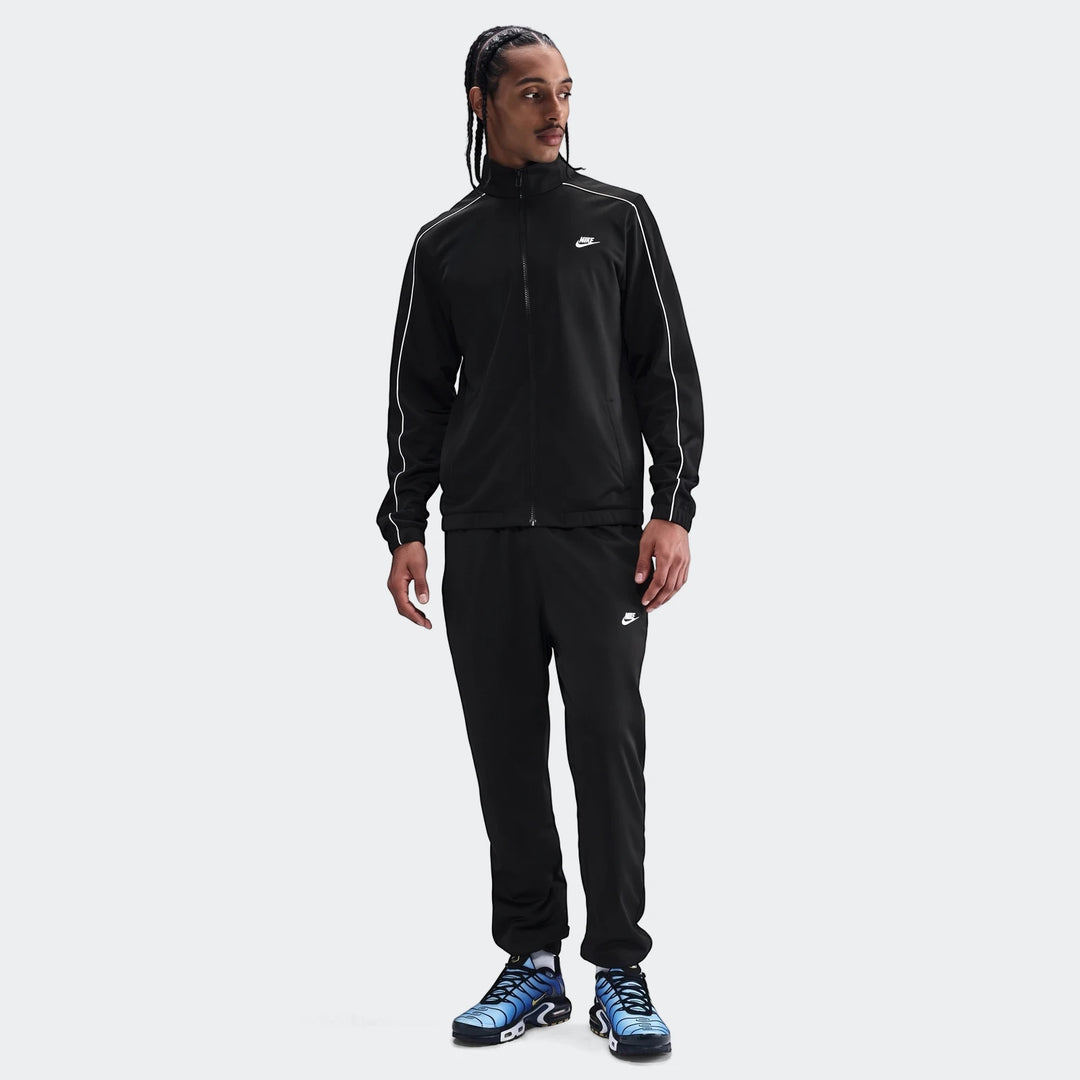 A male model wearing a black Nike Sportswear Club poly-knit tracksuit with white piping, paired with blue and black