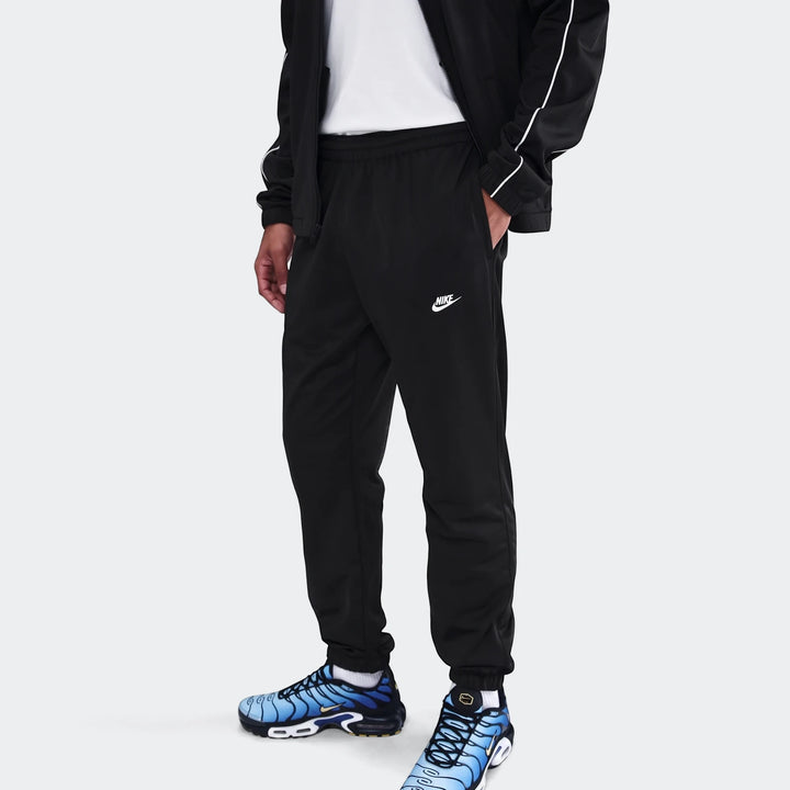 A man wears a black Nike Sportswear Club poly-knit tracksuit with white piping and the Nike swoosh logo on the pants. He