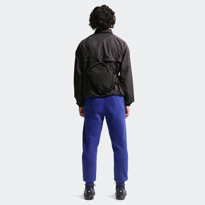 A man from behind wearing royal blue Nike Sportswear Club men's joggers, a dark gray jacket, and sneakers.