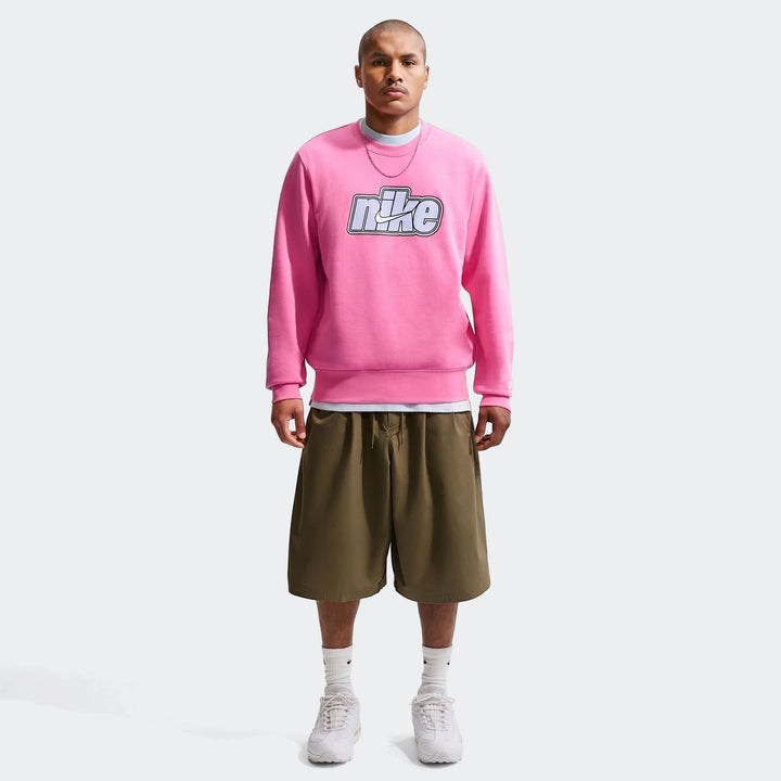 Full body shot of a man wearing a pink Nike Sportswear Club French Terry Crew-Neck Sweatshirt and olive green shorts.