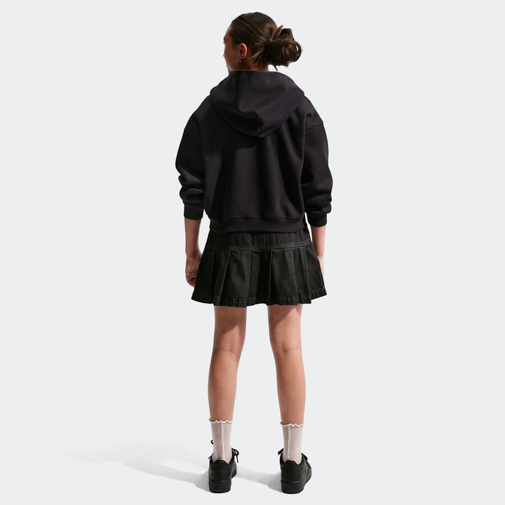 A young girl from the back wearing a black Nike Sportswear Big Kids' denim pleated skirt, a black hoodie, white socks, and