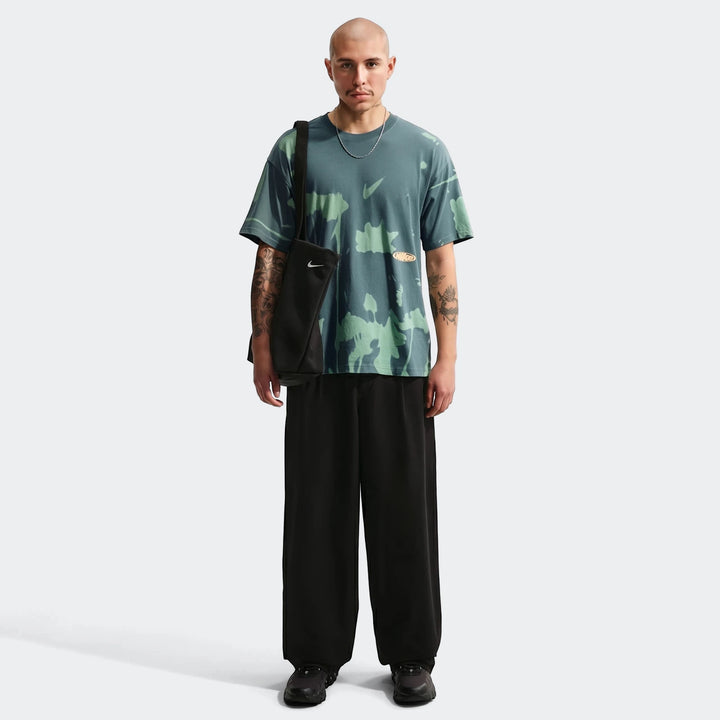 Male model wearing an oversized Nike Sportswear green t-shirt with an abstract pattern, black wide-leg pants, and a black