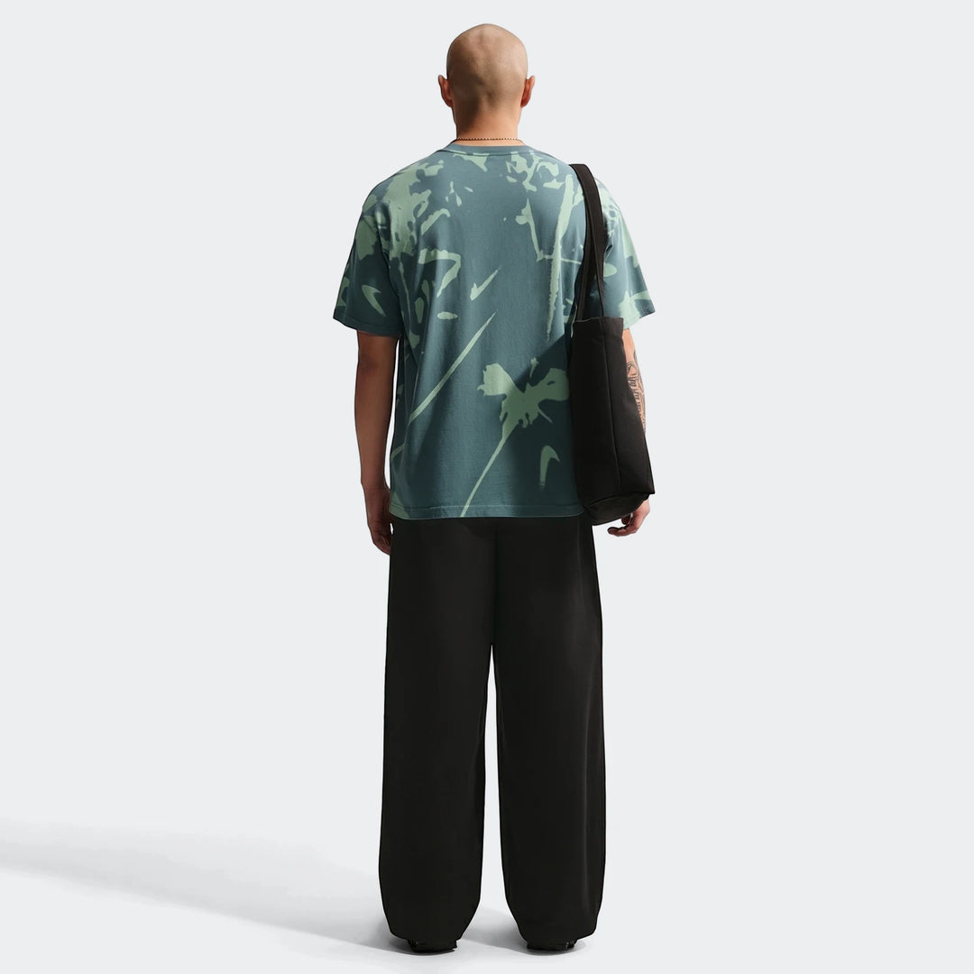 Bald man wearing a Nike Sportswear green t-shirt with an abstract pattern and black wide-leg pants, seen from the back.
