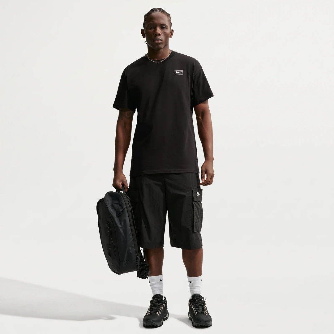 Nike Sportswear men's black t-shirt on a model wearing black cargo shorts, white socks, and black shoes, holding a backpack.