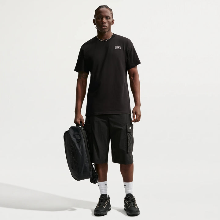 Nike Sportswear men's black t-shirt on a model wearing black cargo shorts, white socks, and black shoes, holding a backpack.