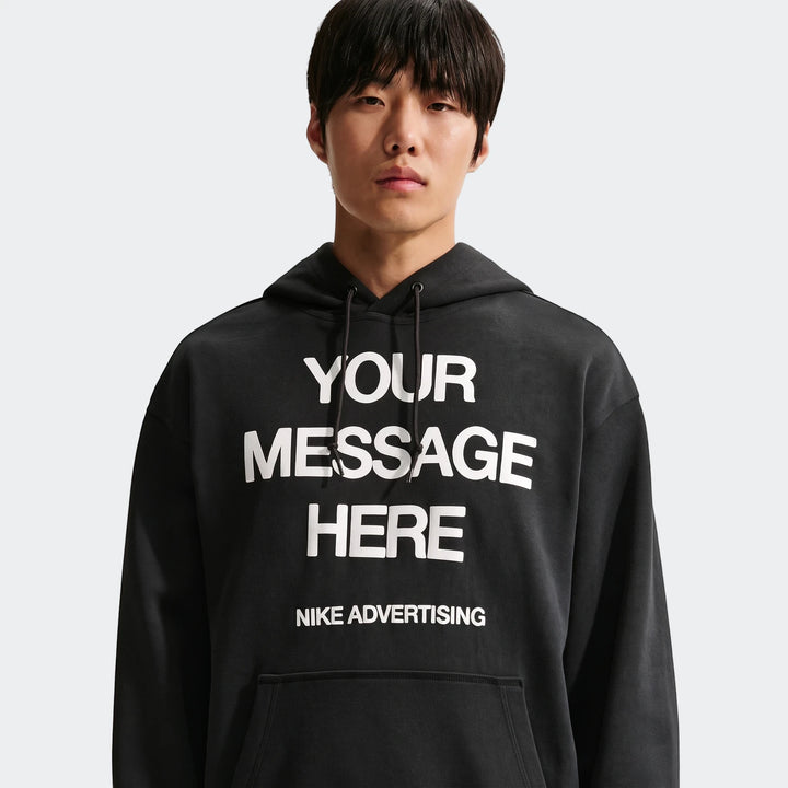 Male model wearing a black Nike Sportswear fleece pullover hoodie with 'Your Message Here' graphic print.