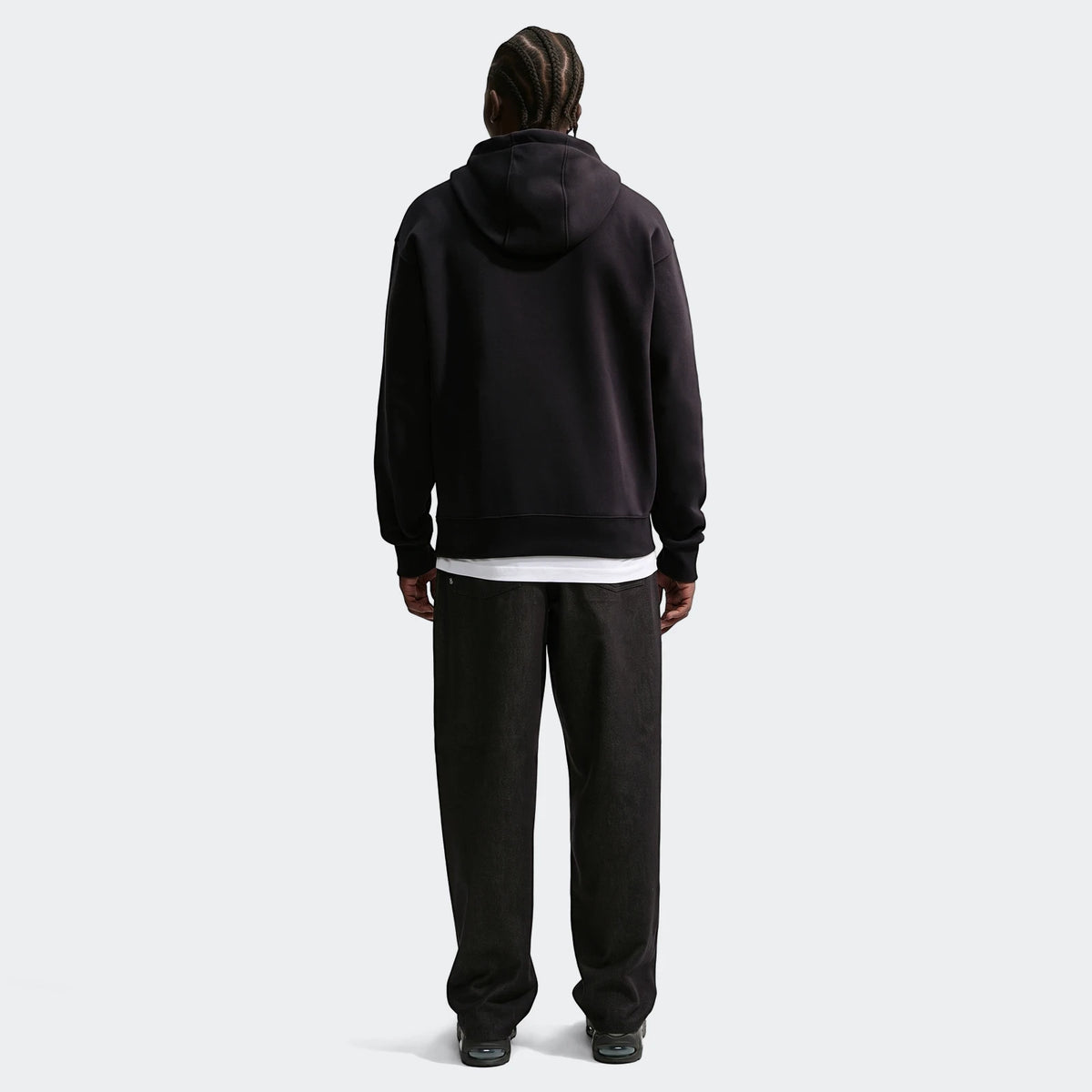 Back view of a man wearing a black Nike Sportswear pullover hoodie and dark wide-leg pants on an off-white background.