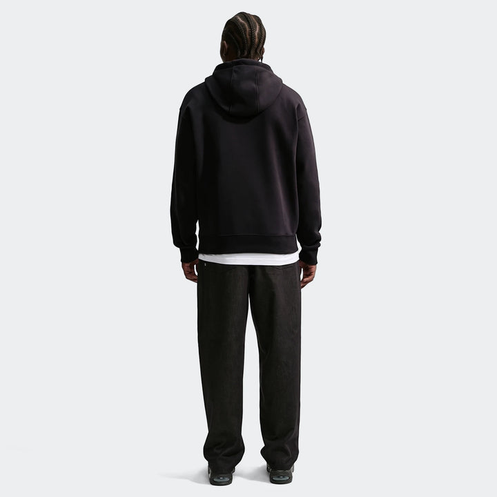 Back view of a man wearing a black Nike Sportswear pullover hoodie and dark wide-leg pants on an off-white background.