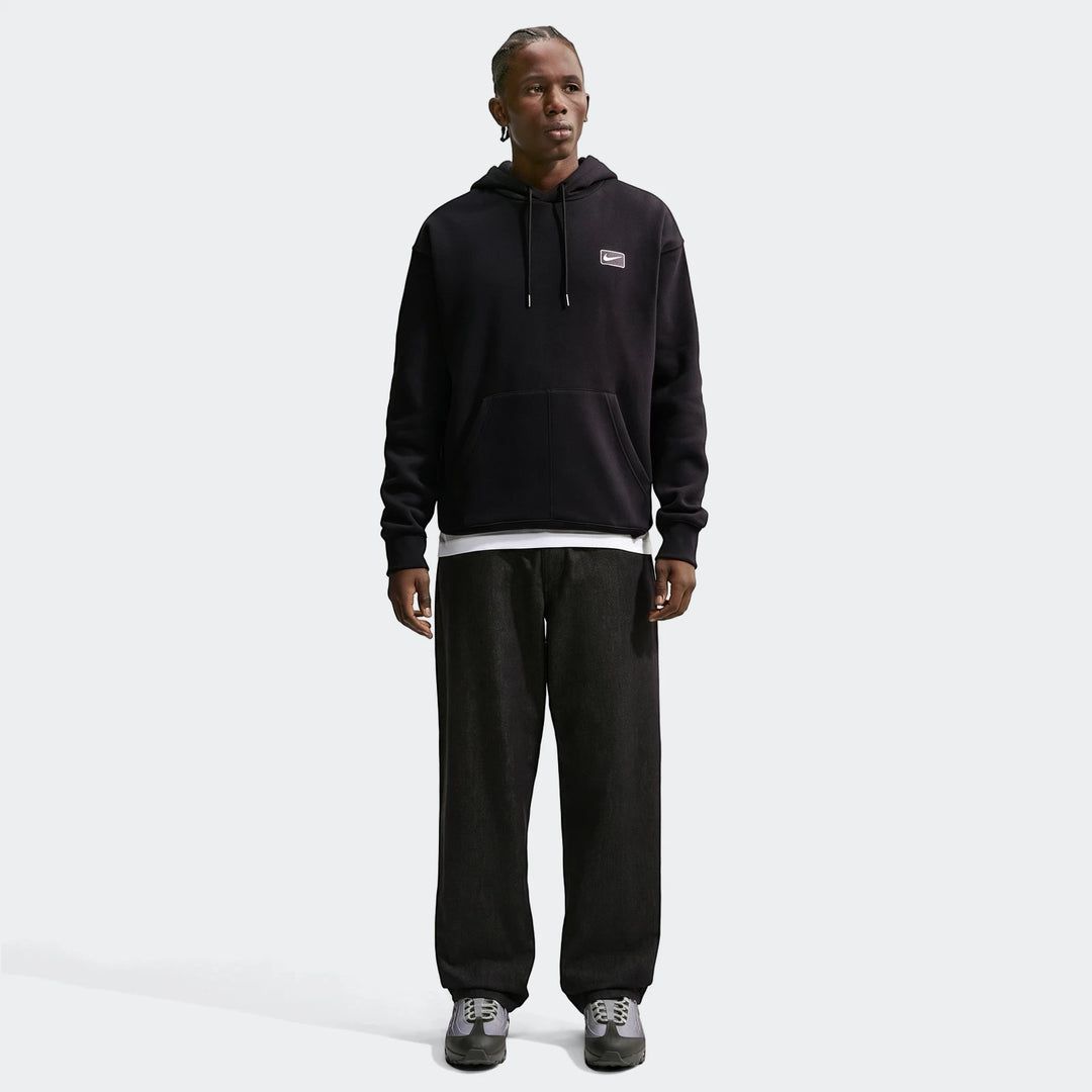 A male model with dreadlocks wears a black Nike Sportswear Men's Pullover Hoodie with dark wide-leg pants and sneakers.