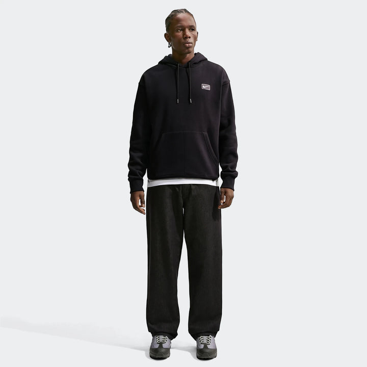 A male model with dreadlocks wears a black Nike Sportswear Men's Pullover Hoodie with dark wide-leg pants and sneakers.