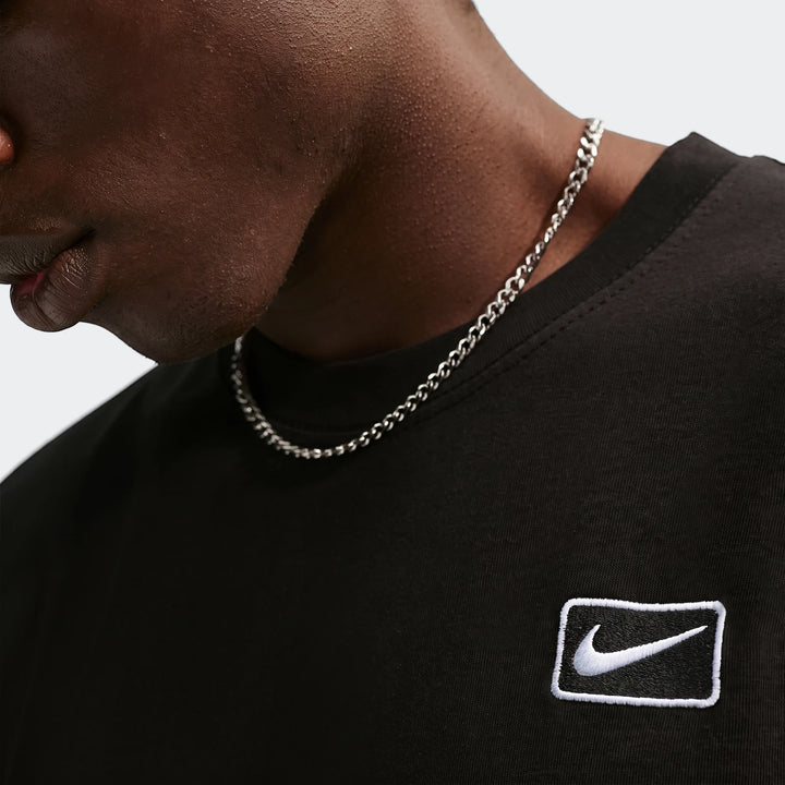 Close-up of a man wearing a black Nike Sportswear Men's T-shirt featuring a white embroidered swoosh logo and a silver chain.
