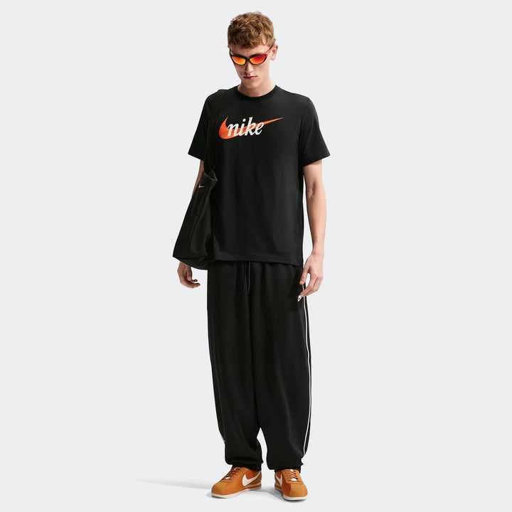 A male model wears a black Nike Sportswear men's t-shirt, black pants with white stripes, and orange shoes with a tote bag.