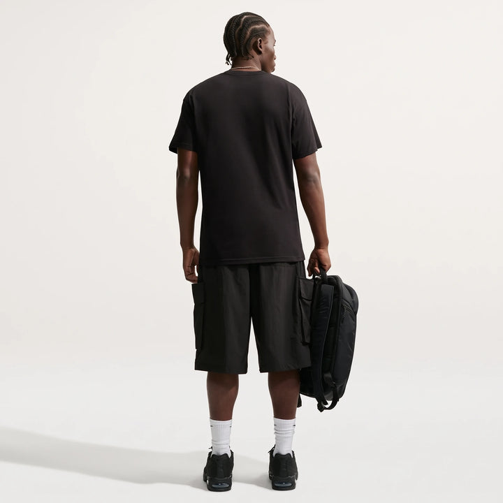 Rear view of a man wearing a black Nike Sportswear men's t-shirt, black cargo shorts, and sneakers, holding a black backpack.
