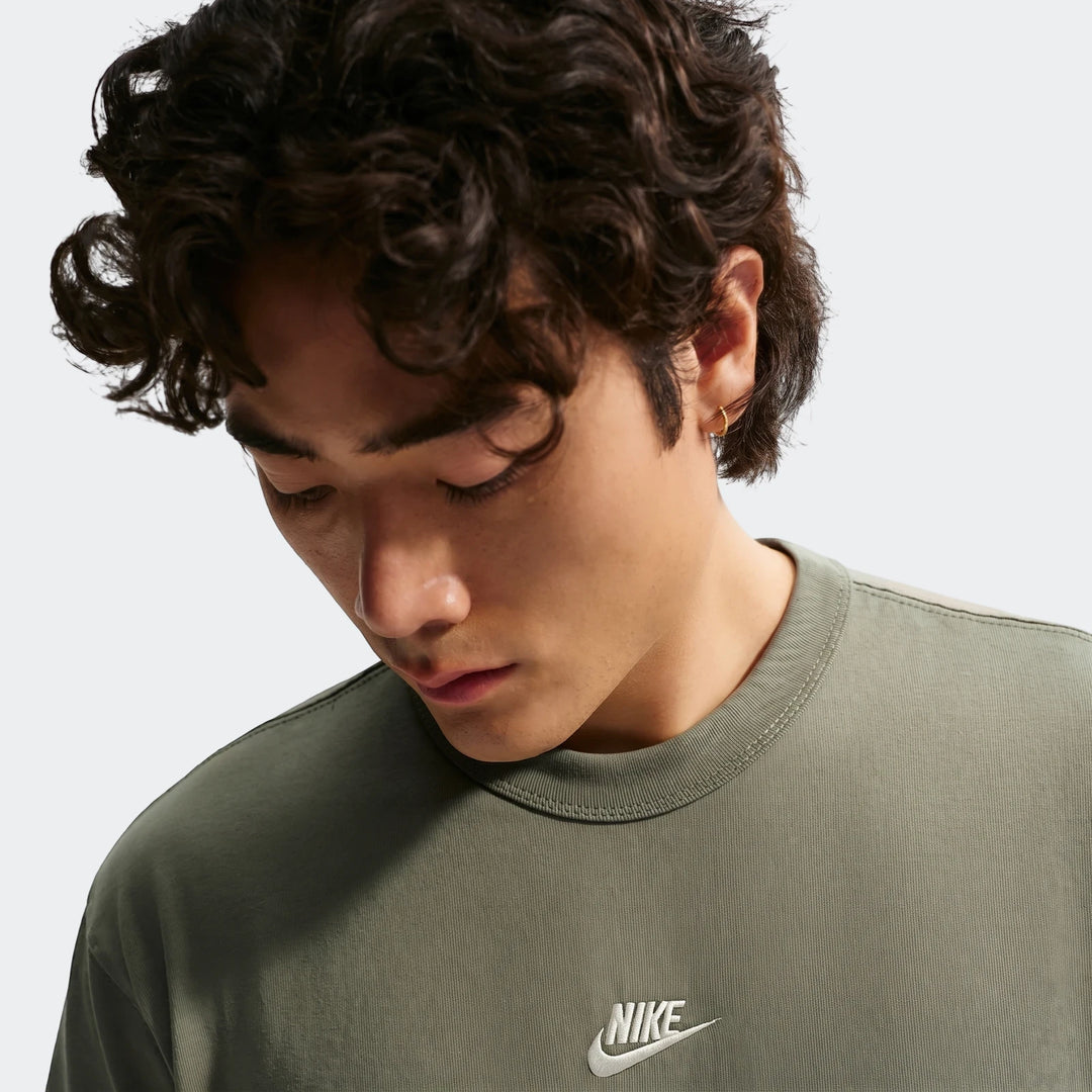 Close-up of a man wearing a green Nike Sportswear Premium Essentials men's t-shirt with a white Nike logo. The man has curly
