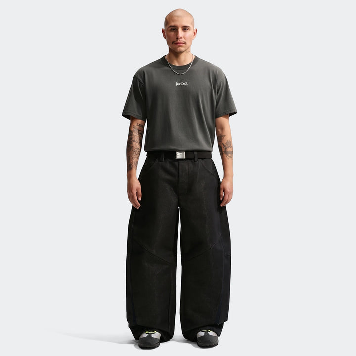 A bald male model with tattoos wears a dark grey Nike Sportswear Premium Essentials T-Shirt and black wide-leg pants.