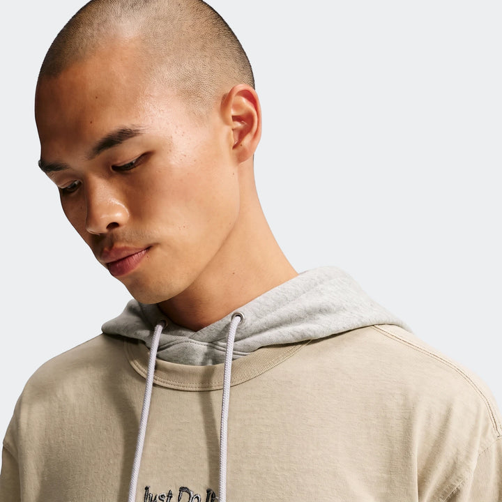 Nike Sportswear Premium Essentials T-Shirt in beige, worn by a man layered over a grey hoodie, looking down.