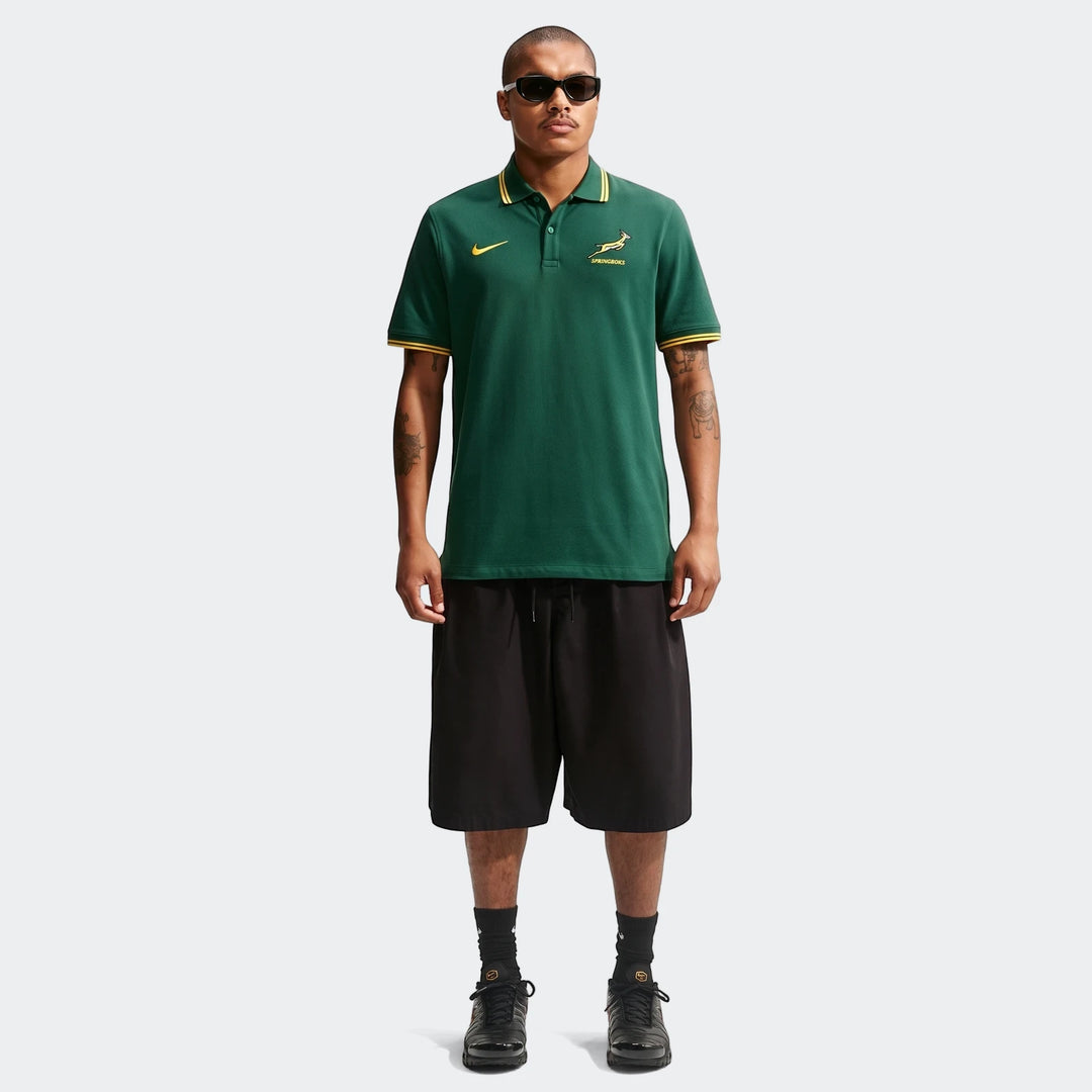 A male model stands wearing a Nike Springboks Pique Polo Golfer for men in green and gold, paired with black shorts and