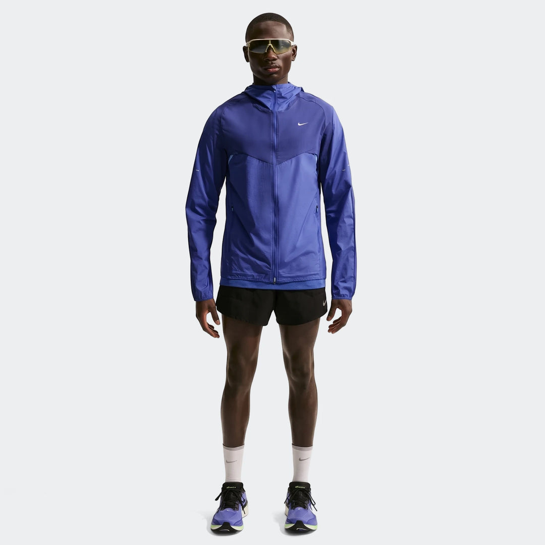Full body shot of a man wearing a Nike Stride Plus Blue Men's Repel Running Jacket, black running shorts, and purple running