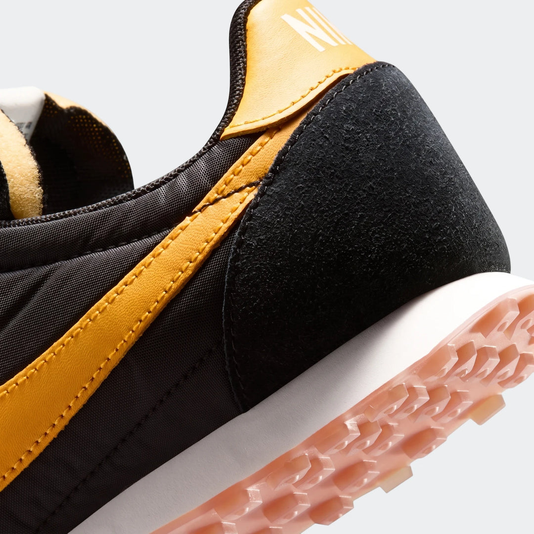 Close-up of a black and gold Nike Waffle Racer men's shoe, detailing the textured heel and prominent gold stripe.