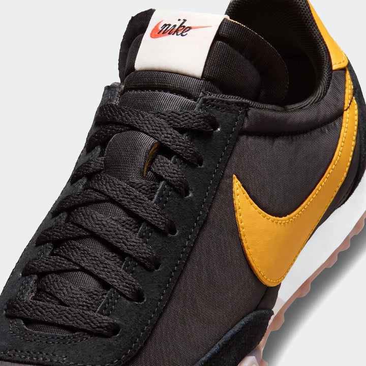 Close-up view of a black Nike Waffle Racer men's shoe featuring a yellow swoosh and black laces.