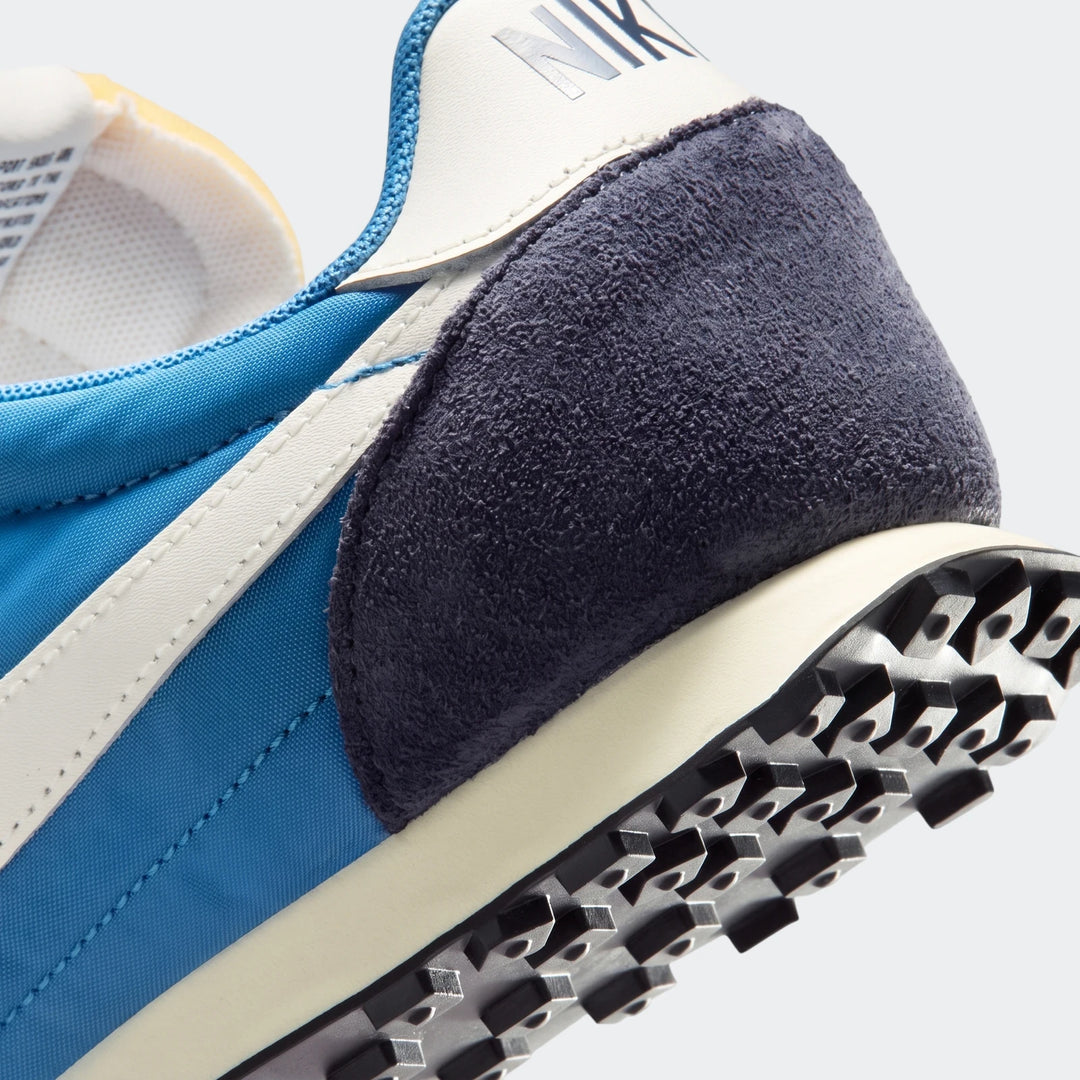 Close-up of a blue and navy suede Nike Waffle Racer Men's shoe, showing the heel, side stripe, and waffle outsole.