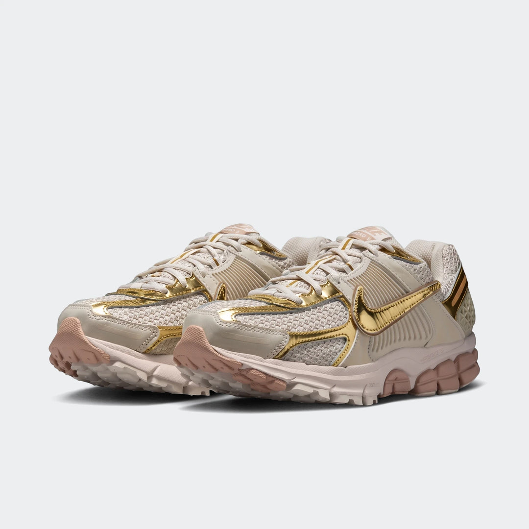 Pair of Nike Zoom Vomero 5 Women's Shoes in beige with shiny gold metallic accents, shown from a low angle.