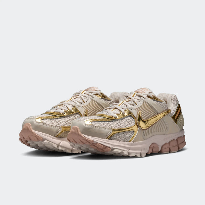 Pair of Nike Zoom Vomero 5 Women's Shoes in beige with shiny gold metallic accents, shown from a low angle.