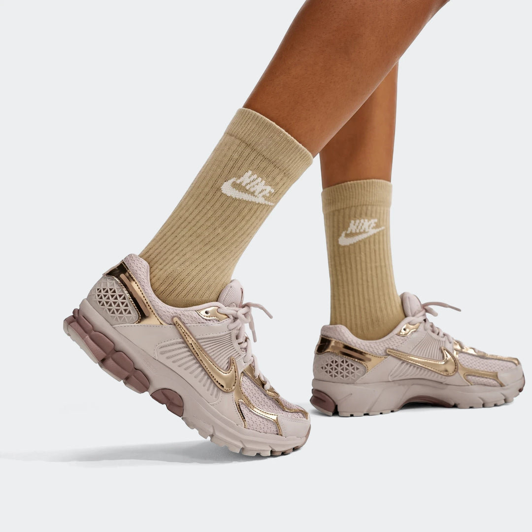 Nike Zoom Vomero 5 women's shoes in light pink with metallic rose gold accents, worn on feet with tan Nike socks.