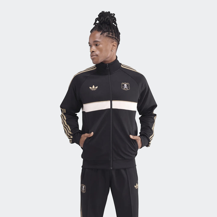 Male model wearing a black and gold Adidas Op Og2 Tt track jacket and matching pants, standing with hands in pockets.