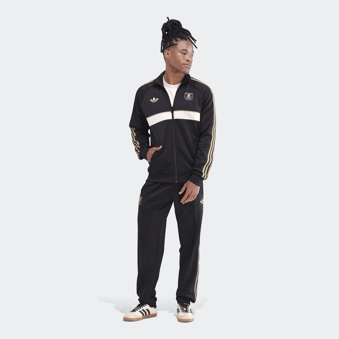 Male model wearing an Op Og2 Tt Adidas tracksuit in black, white, and gold with the Orlando Pirates logo, standing full body.