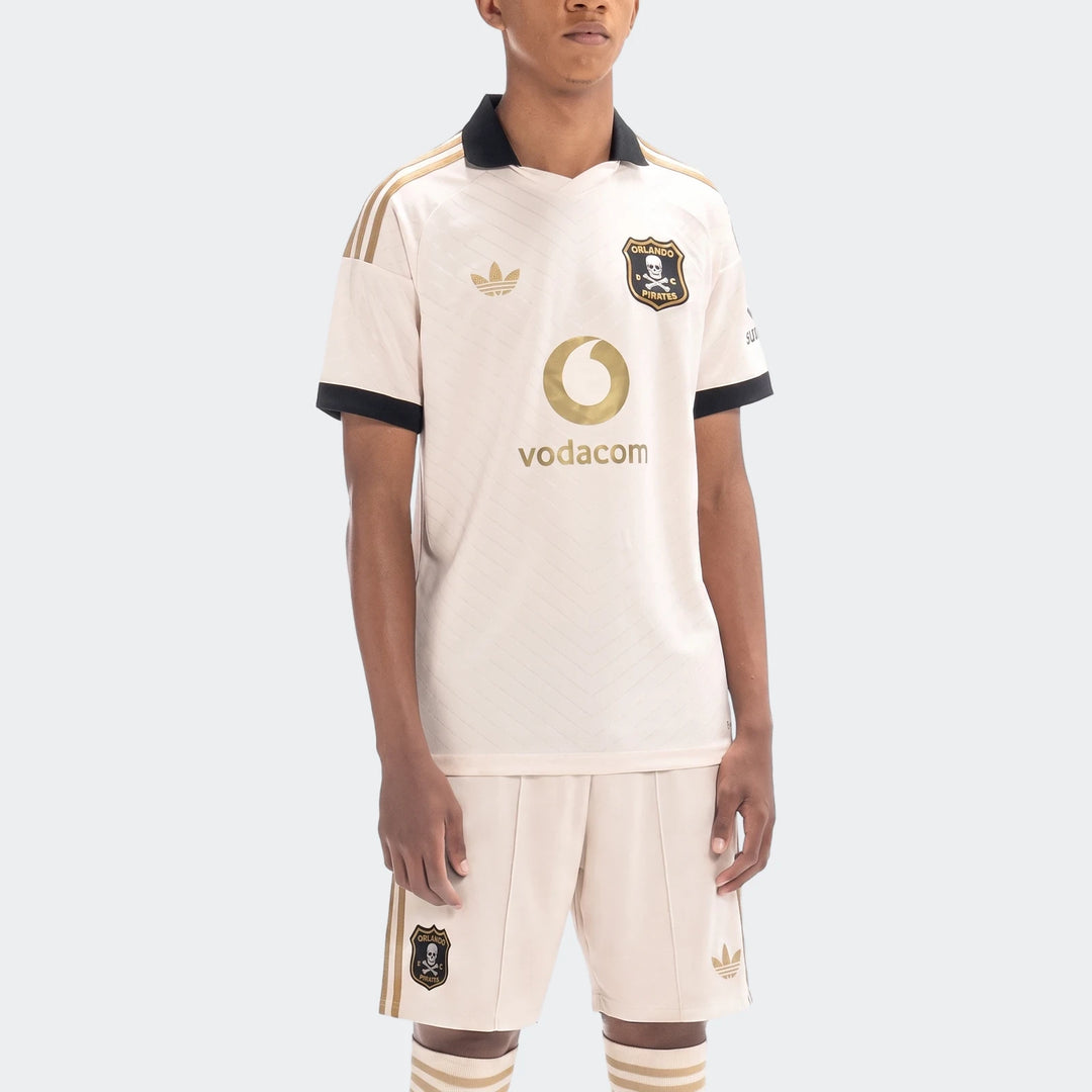 Male model wearing an Orlando Pirates 26/27 Away Kit, featuring beige jersey and shorts with gold Adidas logos and team