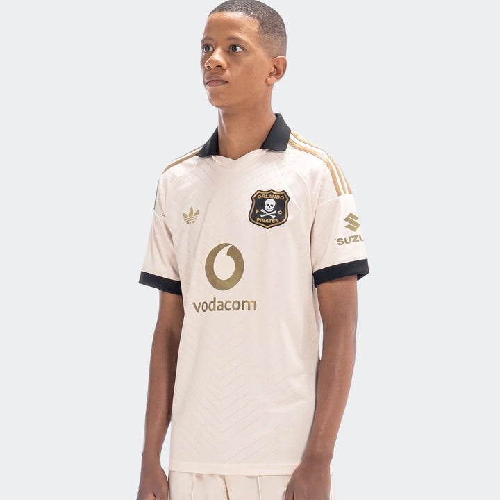 Young male model wearing an Orlando Pirates 26/27 Away Jersey in cream and black with gold logos and details, shown waist-up.