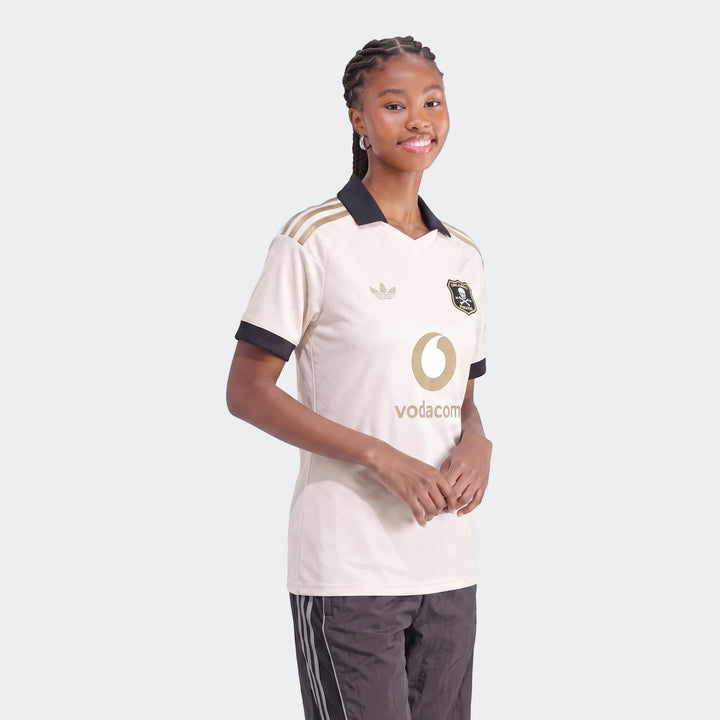 A female model wearing the Orlando Pirates 26/27 Away Jersey, featuring cream, black, and gold details.