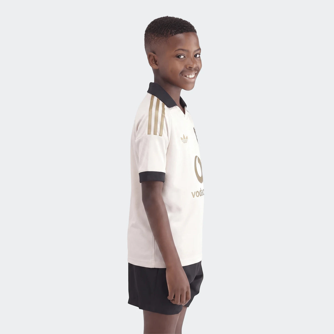 Smiling boy models an Orlando Pirates 26/27 Away Jersey in cream with a black collar and gold details.