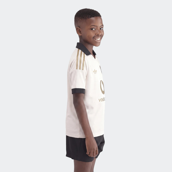Smiling boy models an Orlando Pirates 26/27 Away Jersey in cream with a black collar and gold details.